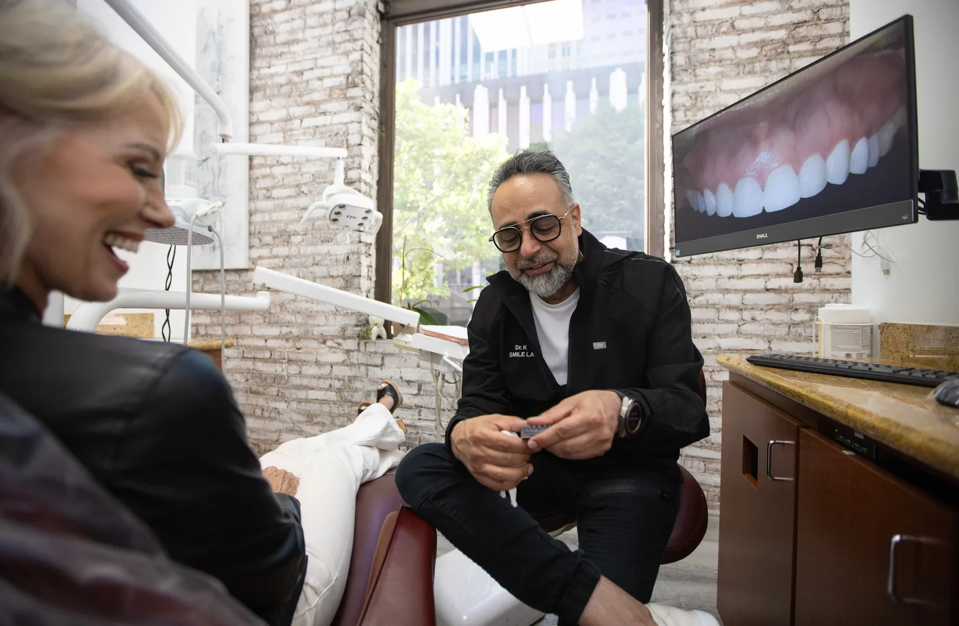 Dr. Kourosh Yousefzadeh seated beside a laughing patient, a digital smile preview on the wall screen