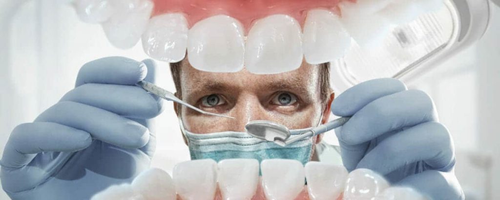 Masked dentist holding instruments, framed by an oversized model of the teeth