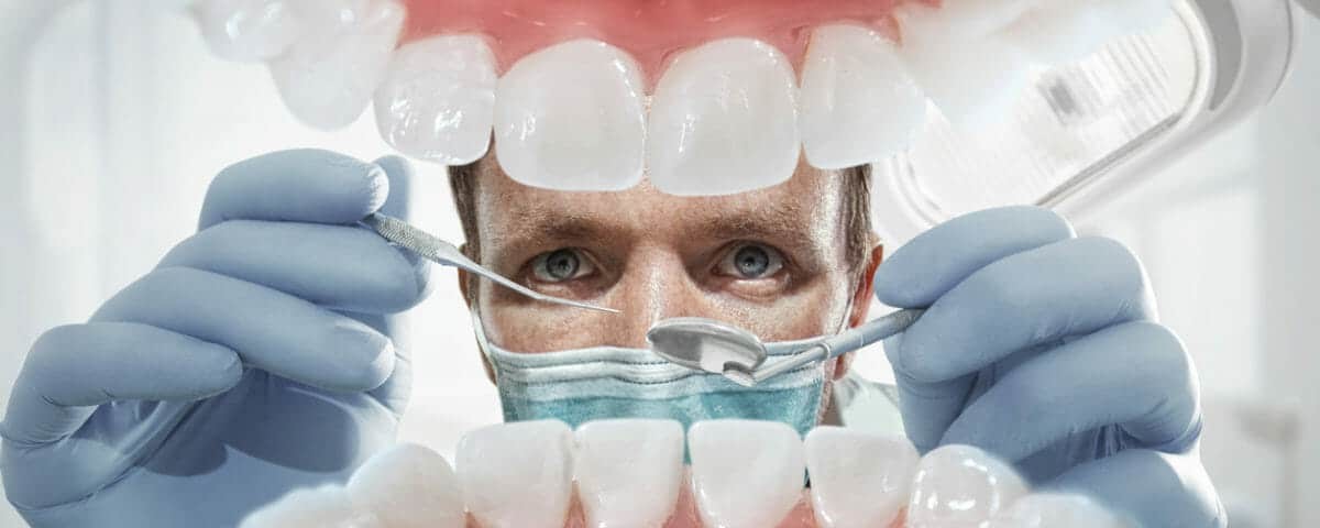Masked dentist holding instruments, framed by an oversized model of the teeth