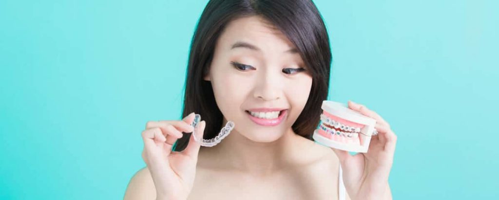 Woman holding a clear aligner in one hand and a model with fixed braces in the other