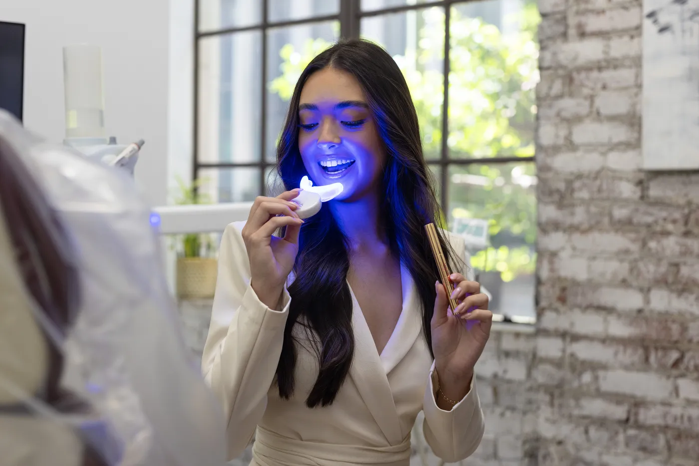 A patient holding a whitening light to her teeth beside the operatory window